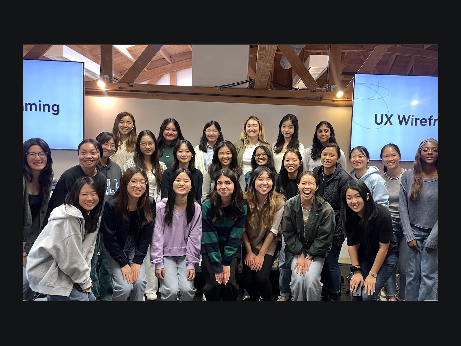A group photo of around twenty five UCLA students with Jennifer at the end of a UX wireframing workshop, standing in front of screens reading "UX Wireframing".